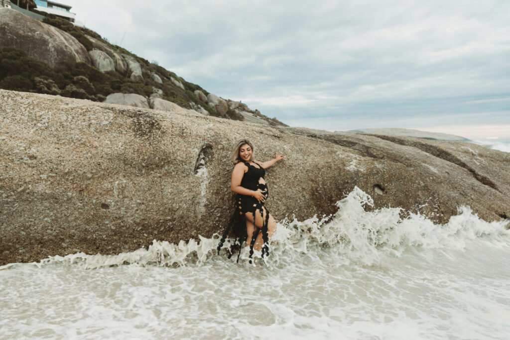 Pregnant woman wearing a black dress posing on Llandudno Beach in Cape Town during a maternity photo session