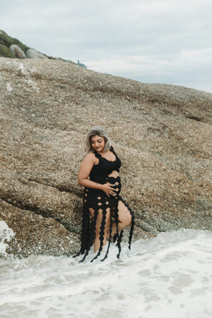 Pregnant woman wearing a black dress posing on Llandudno Beach in Cape Town during a maternity photo session