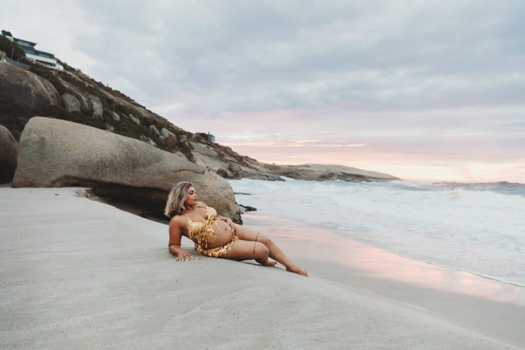 Pregnant woman wearing a gold dress posing on Llandudno Beach sand in Cape Town during a maternity photo session