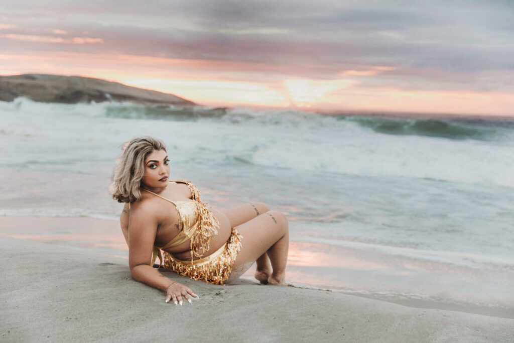 Pregnant woman wearing a gold dress posing on Llandudno Beach sand in Cape Town during a maternity photo session