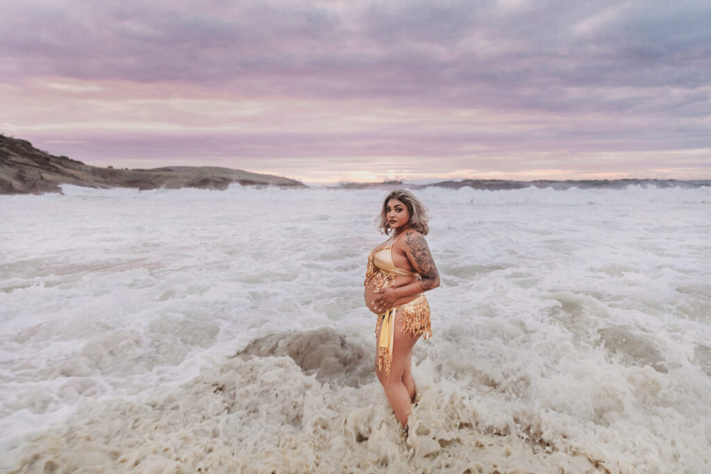 Pregnant woman wearing a gold dress posing on Llandudno Beach sand in Cape Town during a maternity photo session
